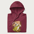 Folded maroon hoodie with cottagecore graphic of a frog under mushrooms with foliage in the background.