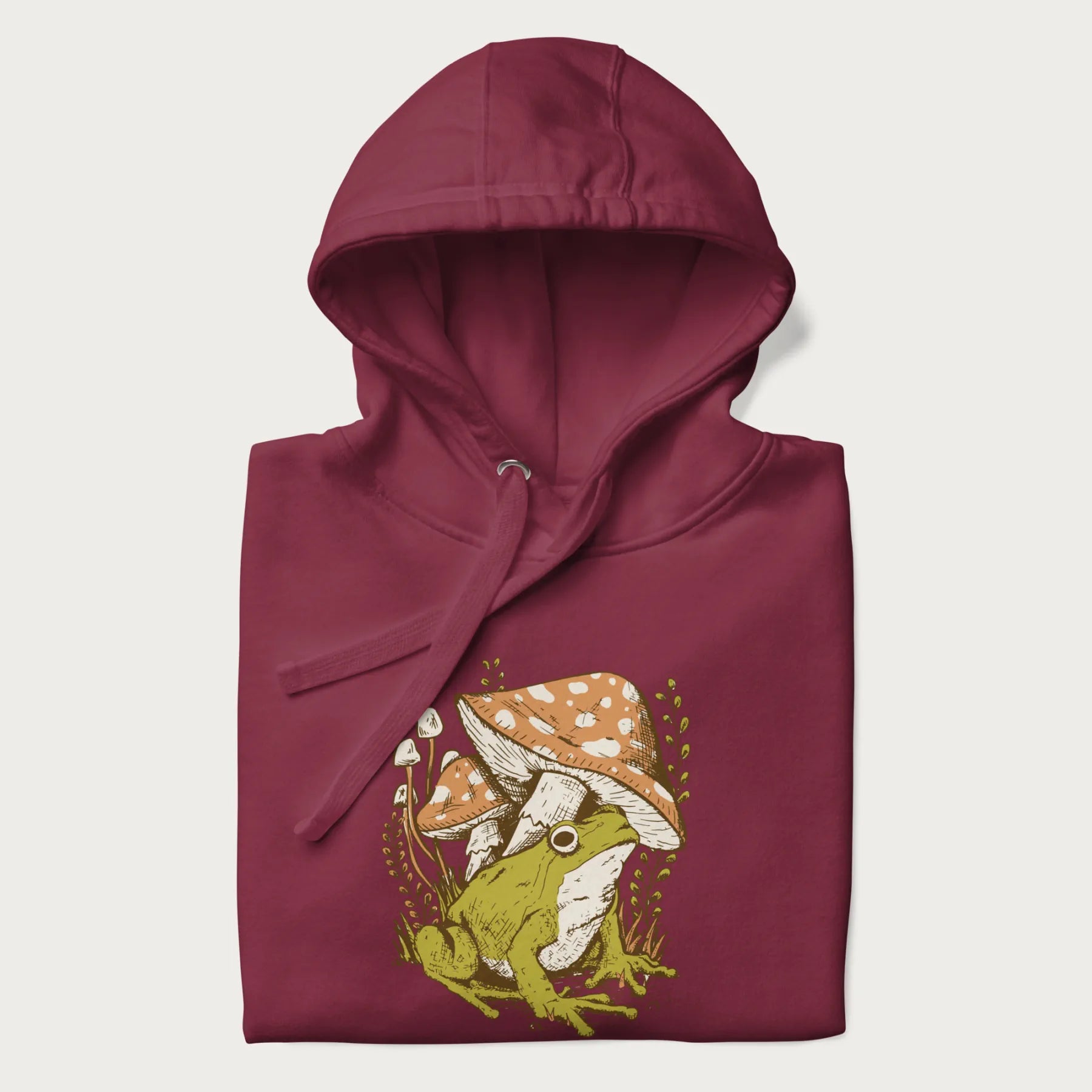 Folded maroon hoodie with cottagecore graphic of a frog under mushrooms with foliage in the background.