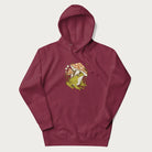 Maroon hoodie with cottagecore graphic of a frog under mushrooms with foliage in the background.