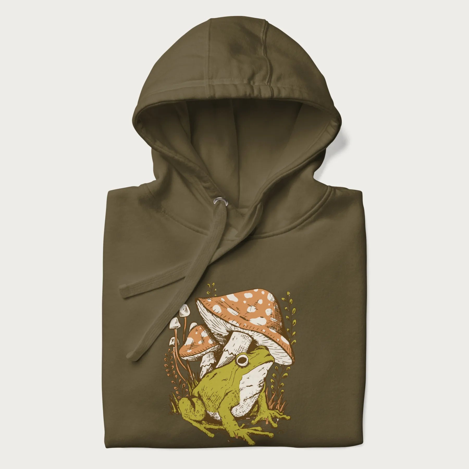 Folded military green hoodie with cottagecore graphic of a frog under mushrooms with foliage in the background.