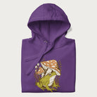 Folded purple hoodie with cottagecore graphic of a frog under mushrooms with foliage in the background.