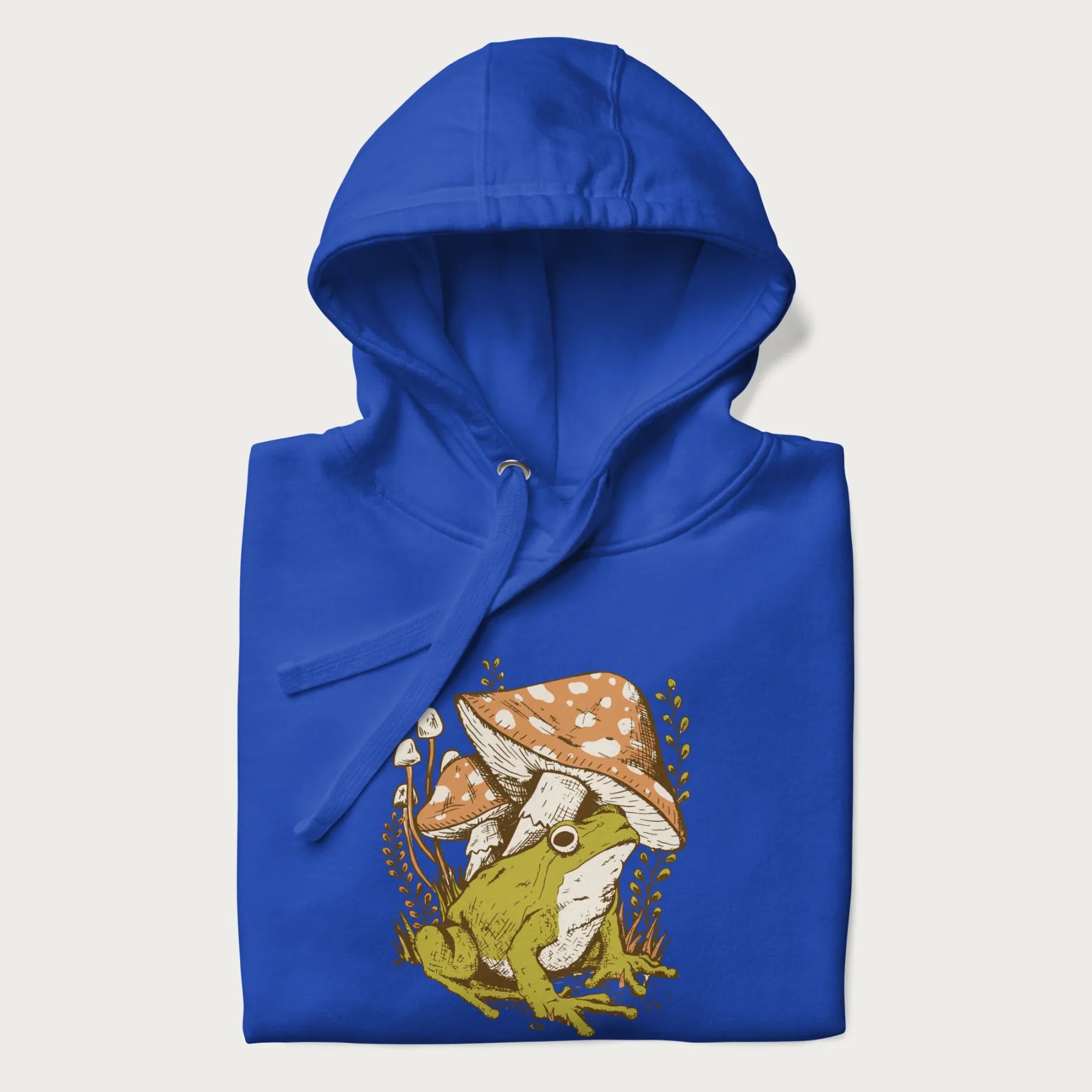 Folded royal blue hoodie with cottagecore graphic of a frog under mushrooms with foliage in the background.