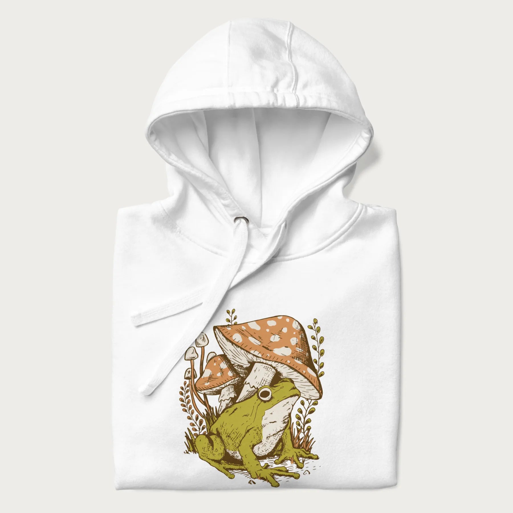 Folded white hoodie with cottagecore graphic of a frog under mushrooms with foliage in the background.