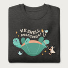 Folded dark grey sweatshirt with a graphic of a turtle flipped on it's shell with the phrase 'We Shell Overcome.'