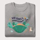 Folded light grey sweatshirt with a graphic of a turtle flipped on it's shell with the phrase 'We Shell Overcome.'